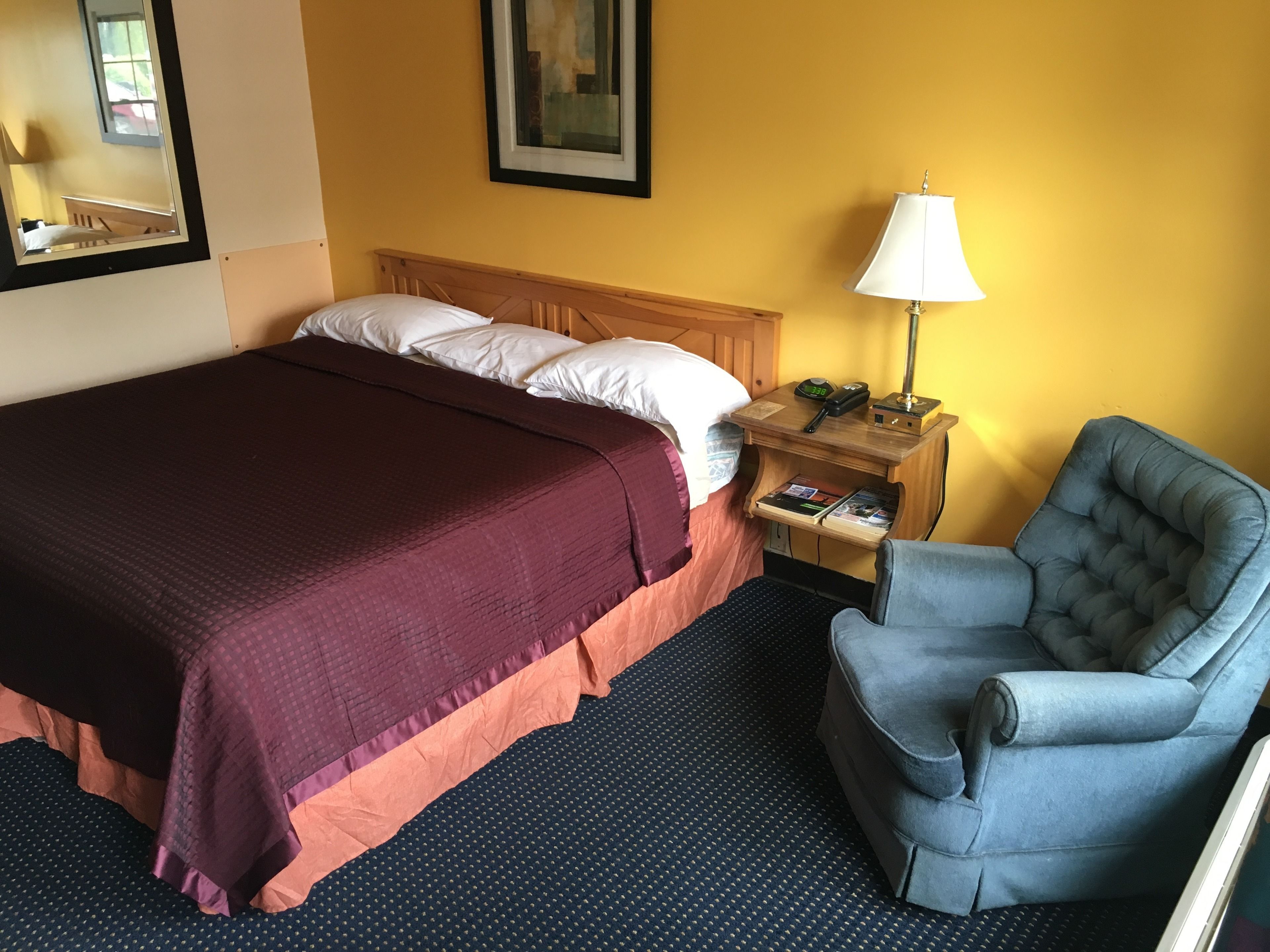 Habitación de hotel con una cama grande, una mesita de noche con lámpara, una silla y un cuadro enmarcado en la pared.
