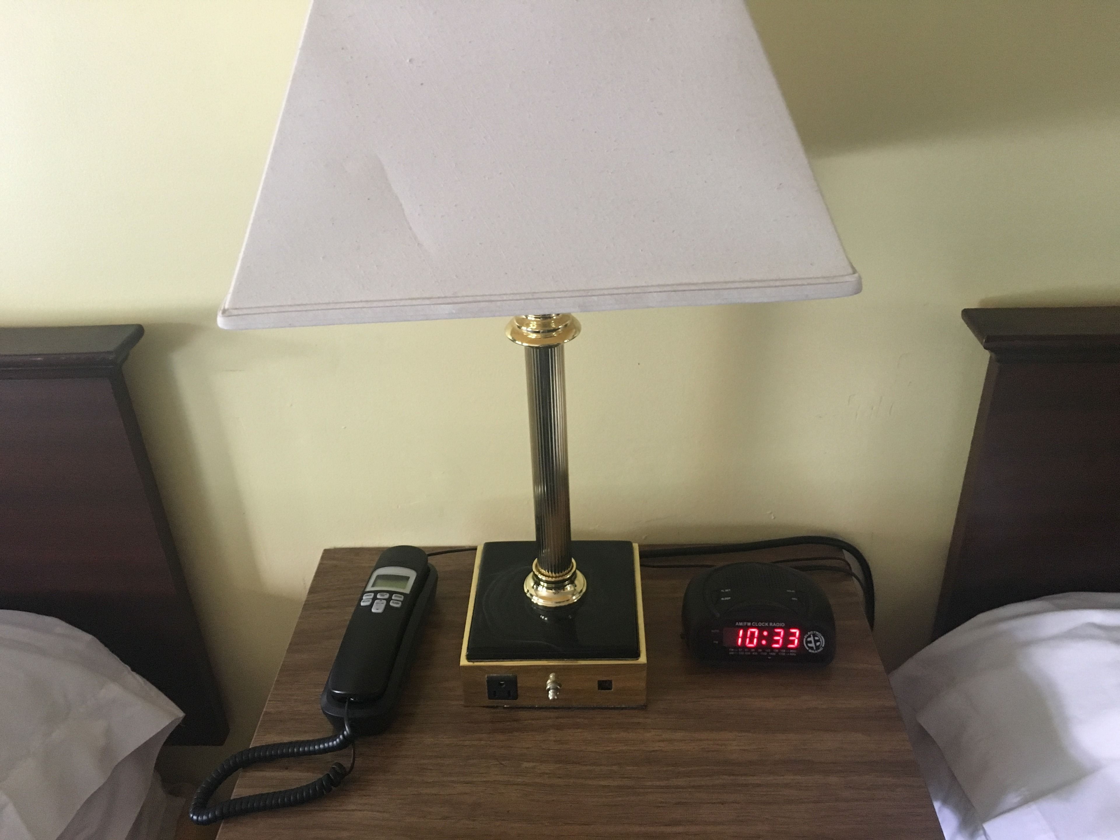 Een nachtkastje met een zwarte telefoon, een radio en een digitale klok waarop 10:33 staat.
