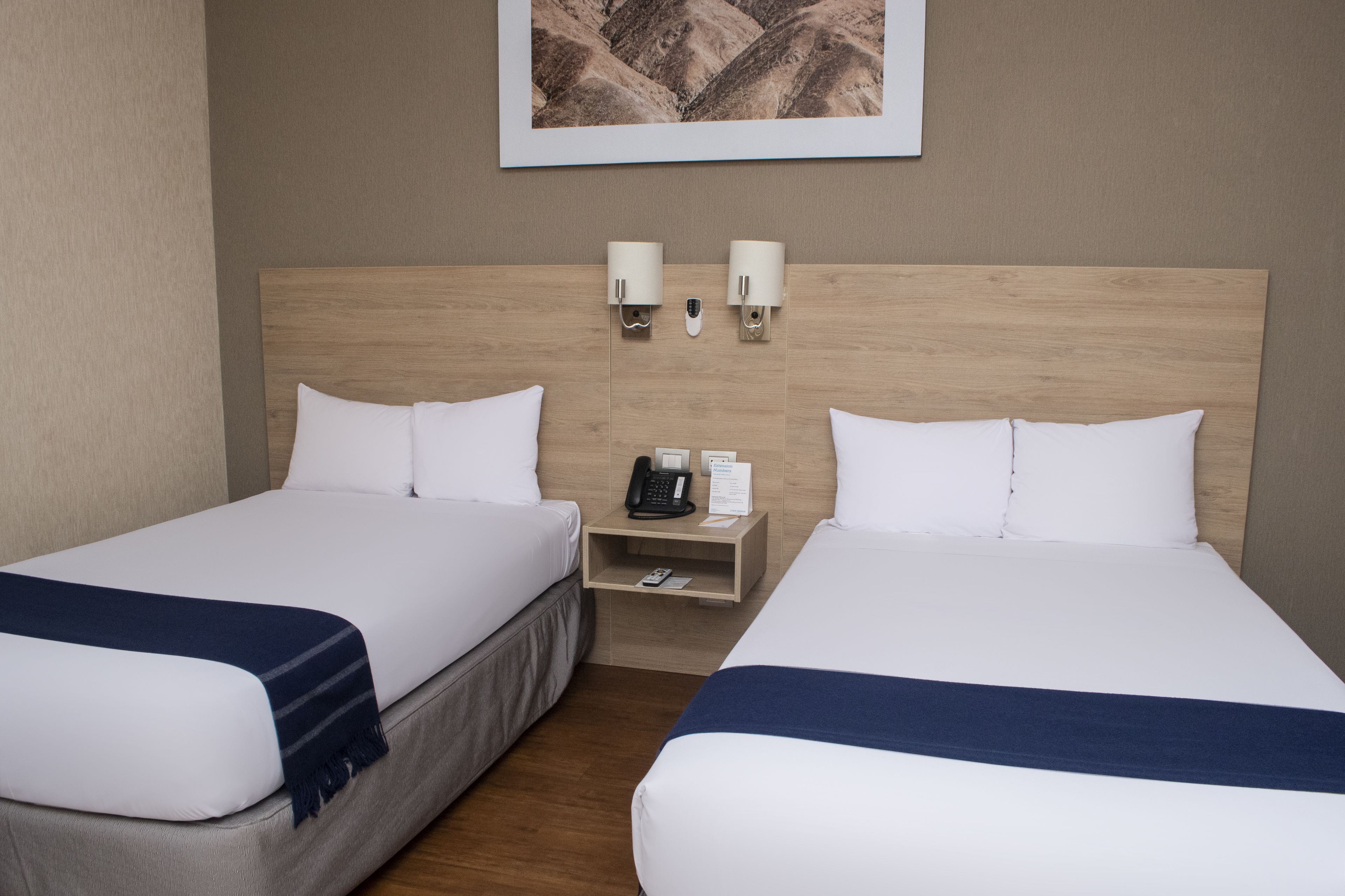 Habitación de hotel con dos camas, cabecera de madera, mesita de noche con teléfono y lámpara, y un cuadro en la pared.