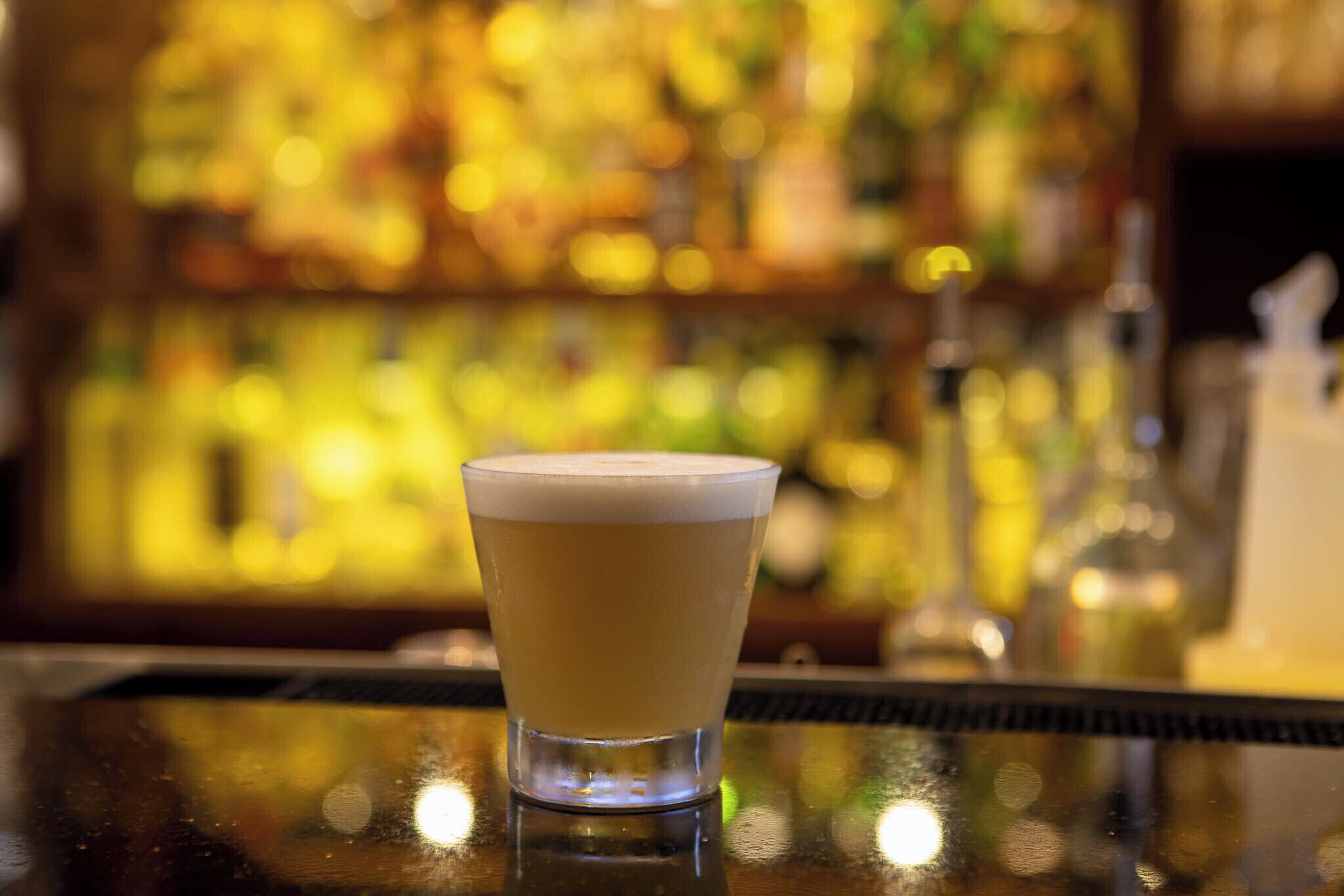 A glass of beer on a bar counter with a blurred background of bottles and a bar.