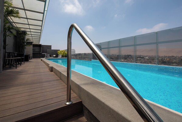 An outdoor swimming pool with a wooden deck and metal railing, overlooking a cityscape.