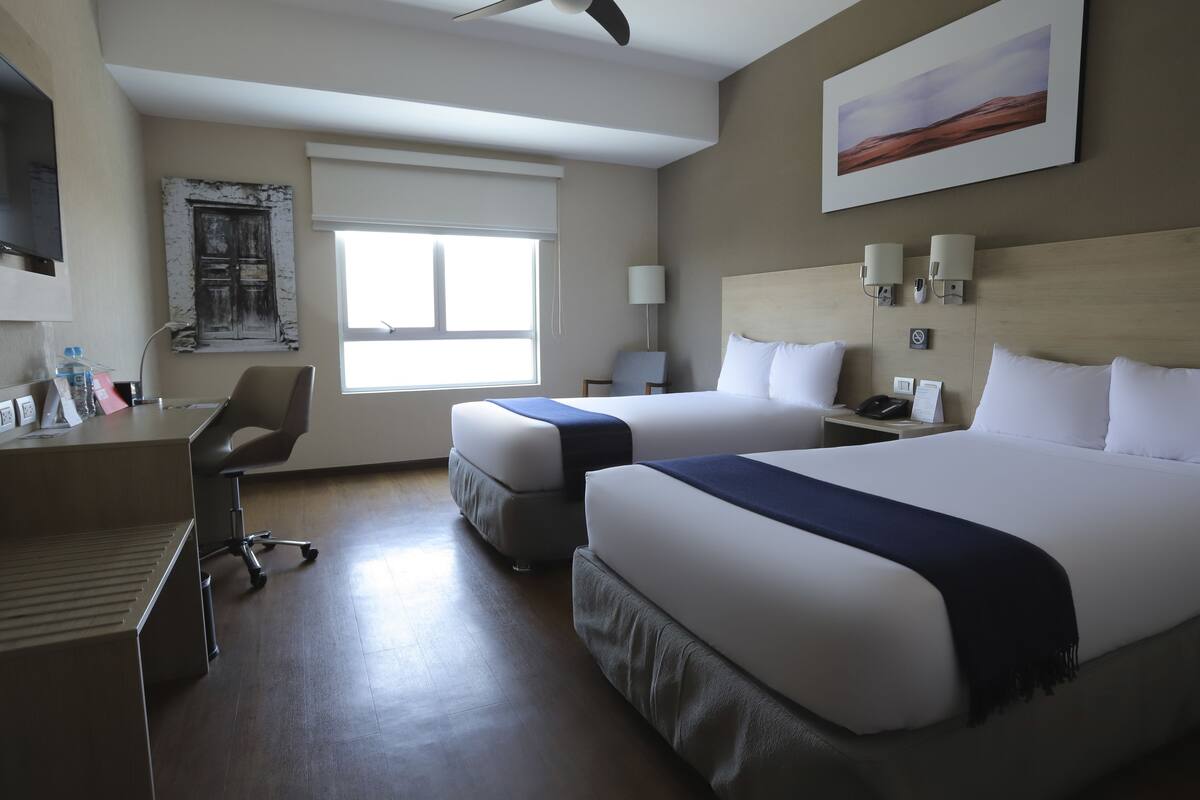 A hotel room with two beds, a desk, a chair, a TV, and a window.