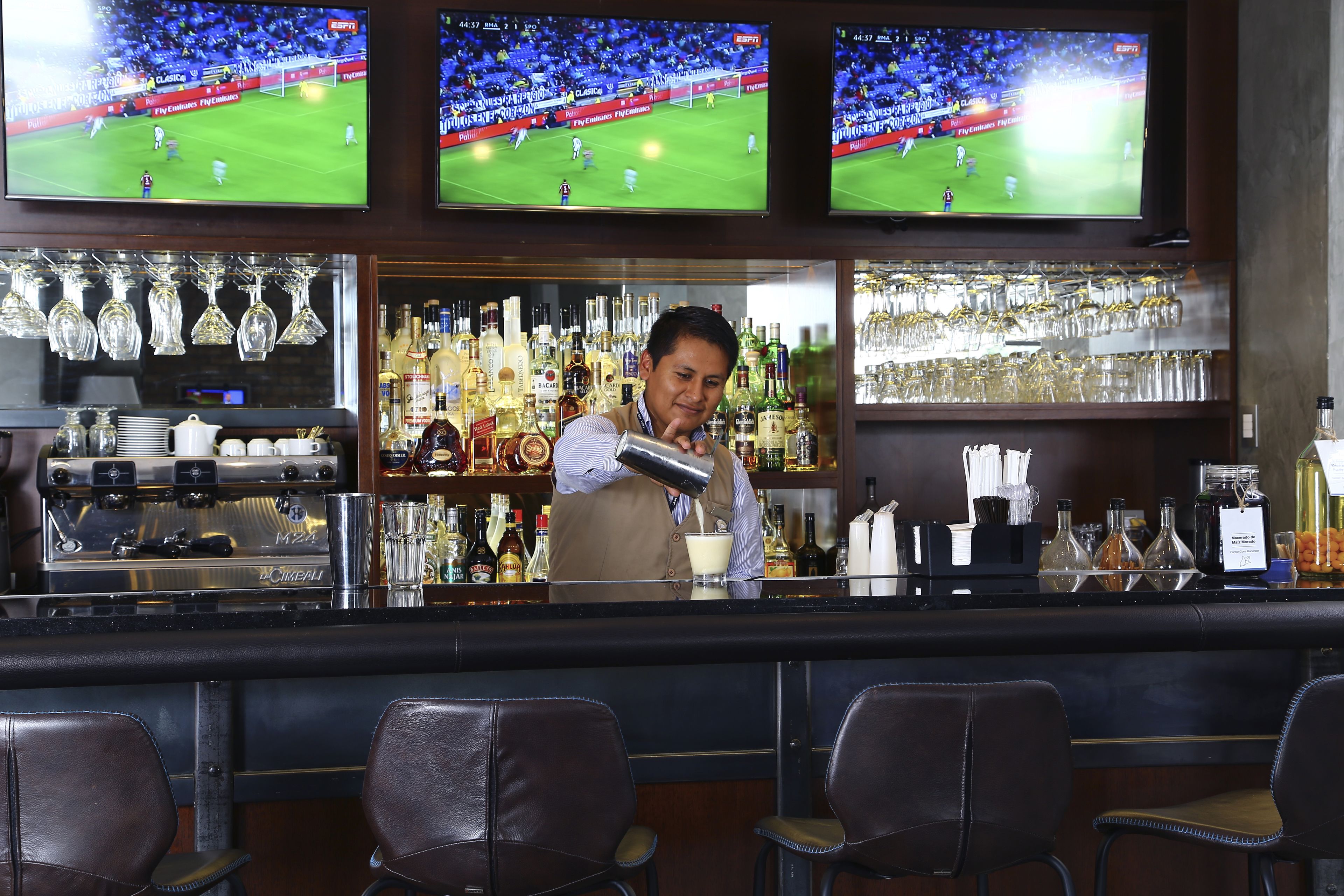 A bar area with multiple screens displaying a soccer game, a barista preparing a drink, and a variety of bottles and glasses on shelves.