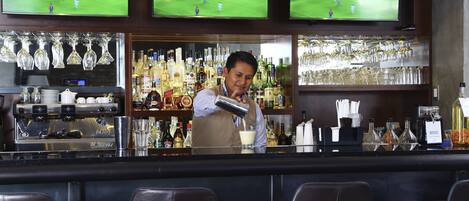 A bar area with multiple screens displaying a soccer game, a barista preparing a drink, and a variety of bottles and glasses on shelves.