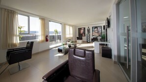 A modern living room with a sofa, a chair, a coffee table, and a TV mounted on the wall. There are large windows with curtains, a small plant on the windowsill, and a view of the cityscape. - Casa Andina Select Tacna (Tacna)