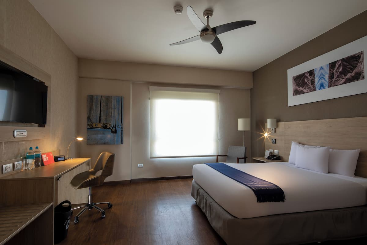 A hotel room with a bed, desk, chair, ceiling fan, artwork, and a window.