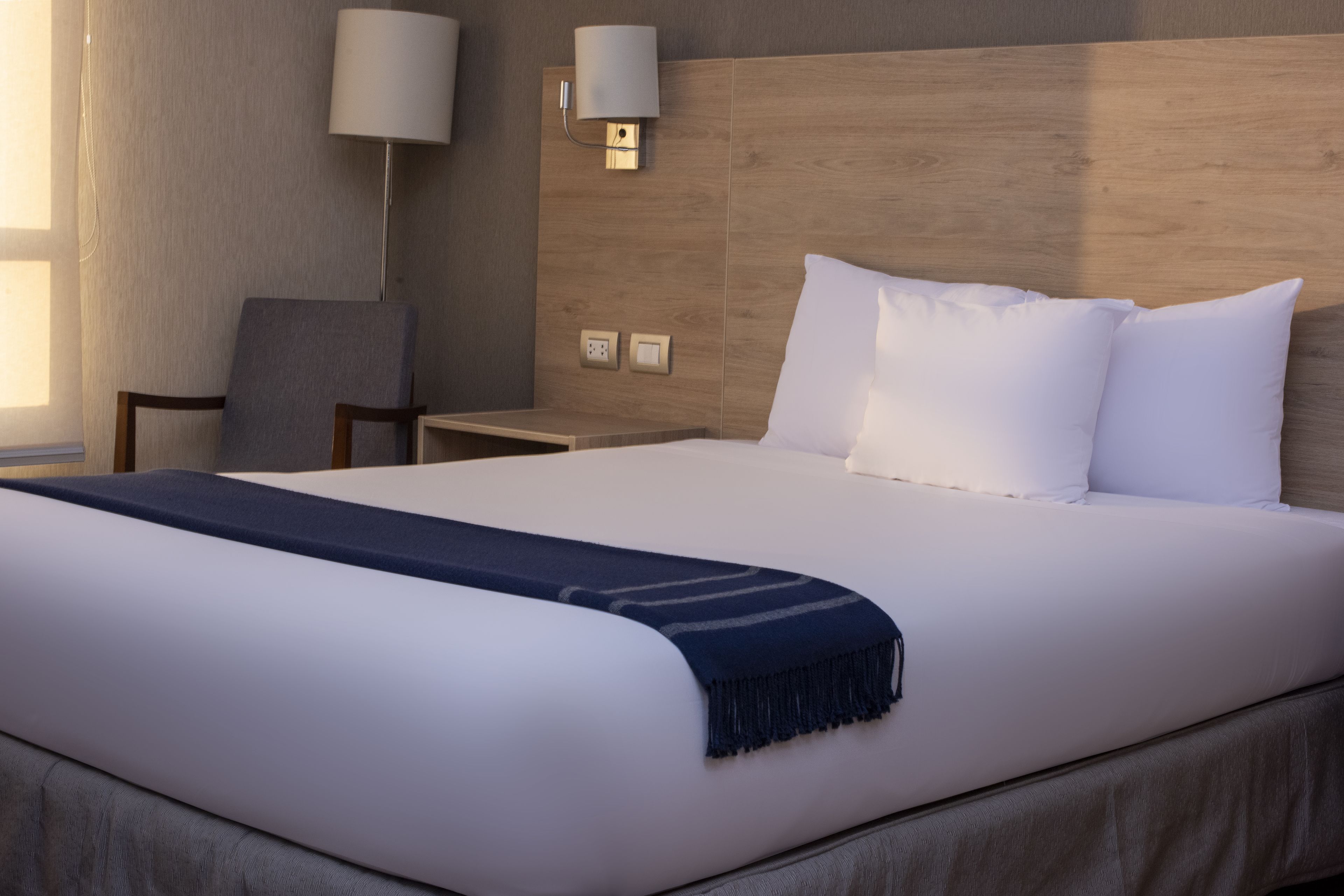Hotellihuone, jossa on suuri sänky, yöpöytä, tuoli ja seinään kiinnitetty lamppu.