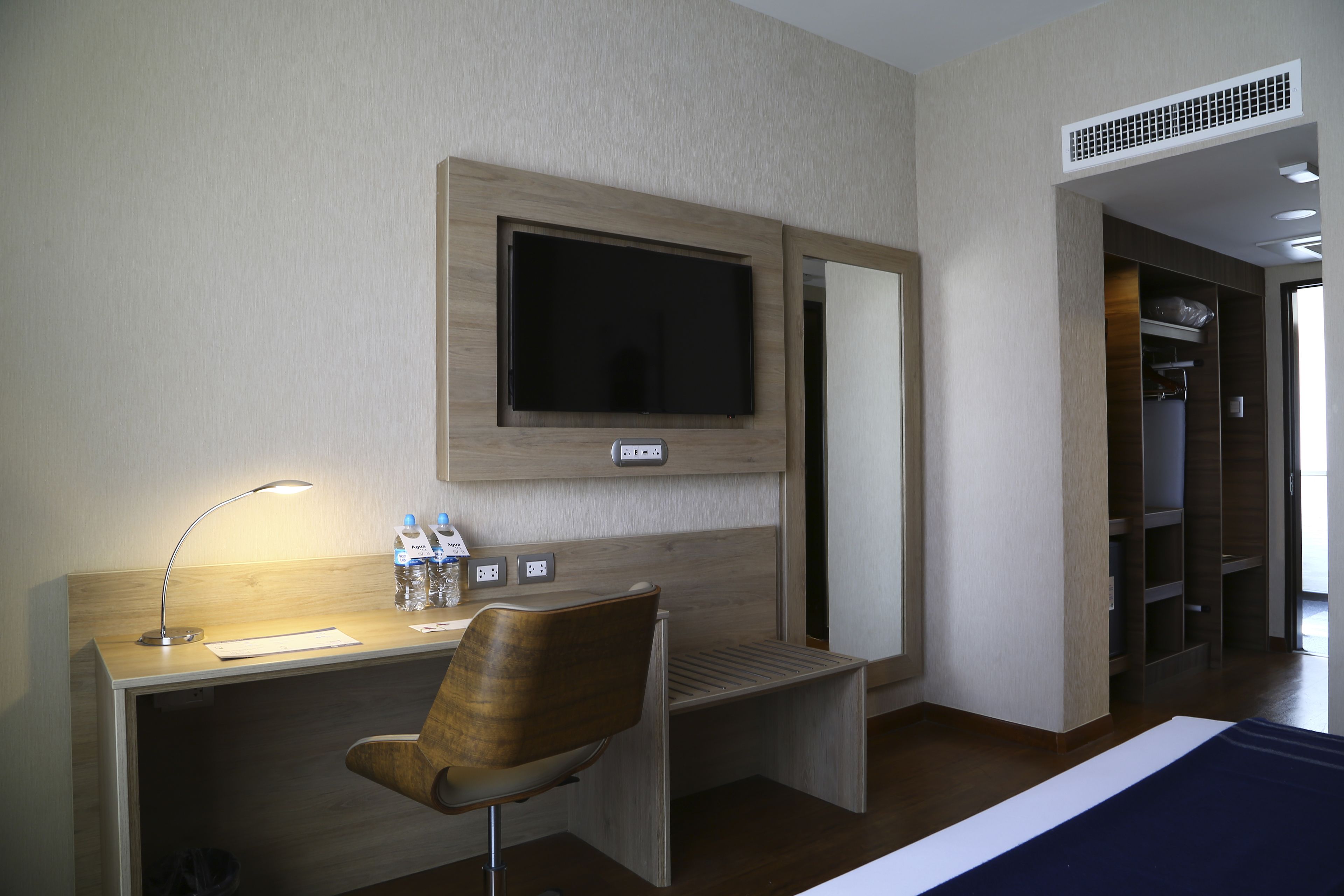 A hotel room with a wooden desk, a chair, a flat-screen TV mounted on the wall, a bedside table, and a walk-in closet.