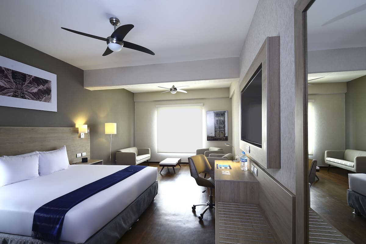 A hotel room with a large bed, a desk with a chair, a ceiling fan, and a large window.