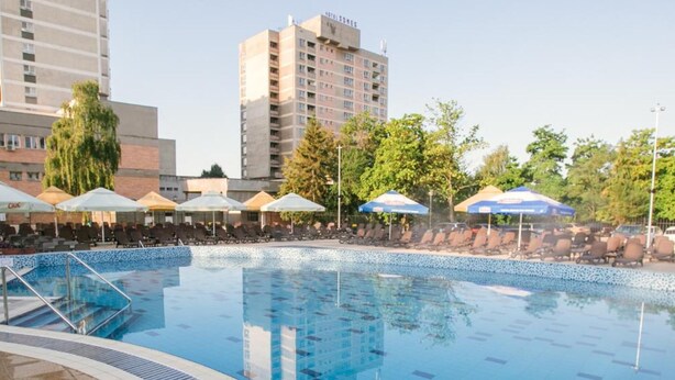 Una piscina con sillones de descanso y sombrillas, rodeada de edificios y árboles.