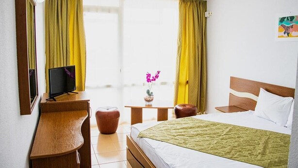 A hotel room with a bed, a desk with a television, a small table with a vase of flowers, and a window with curtains. - Mures Saturn (Saturn)
