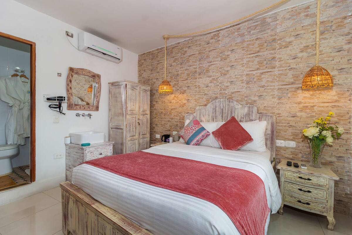 A bedroom with a brick wall, a wooden headboard, a white bed with a red blanket, a bedside table with a vase of flowers, and a hanging light fixture.