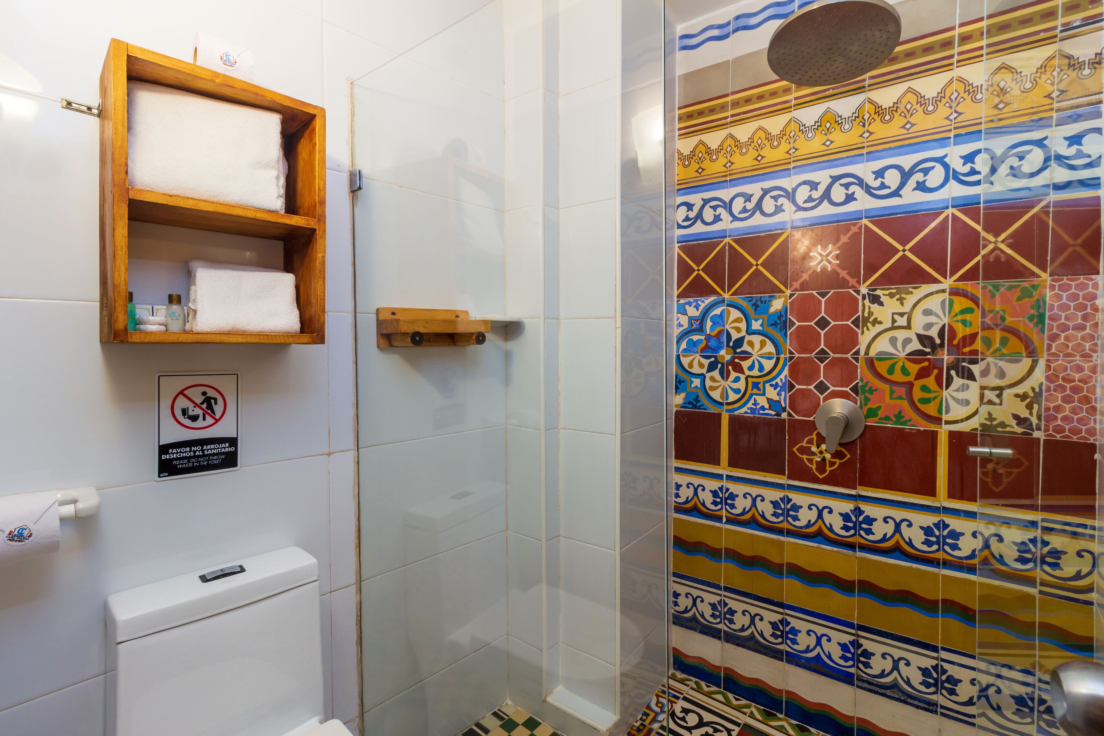 A bathroom with a toilet, a wooden shelf with towels, a shower with a colorful tile wall, and a no-smoking sign.