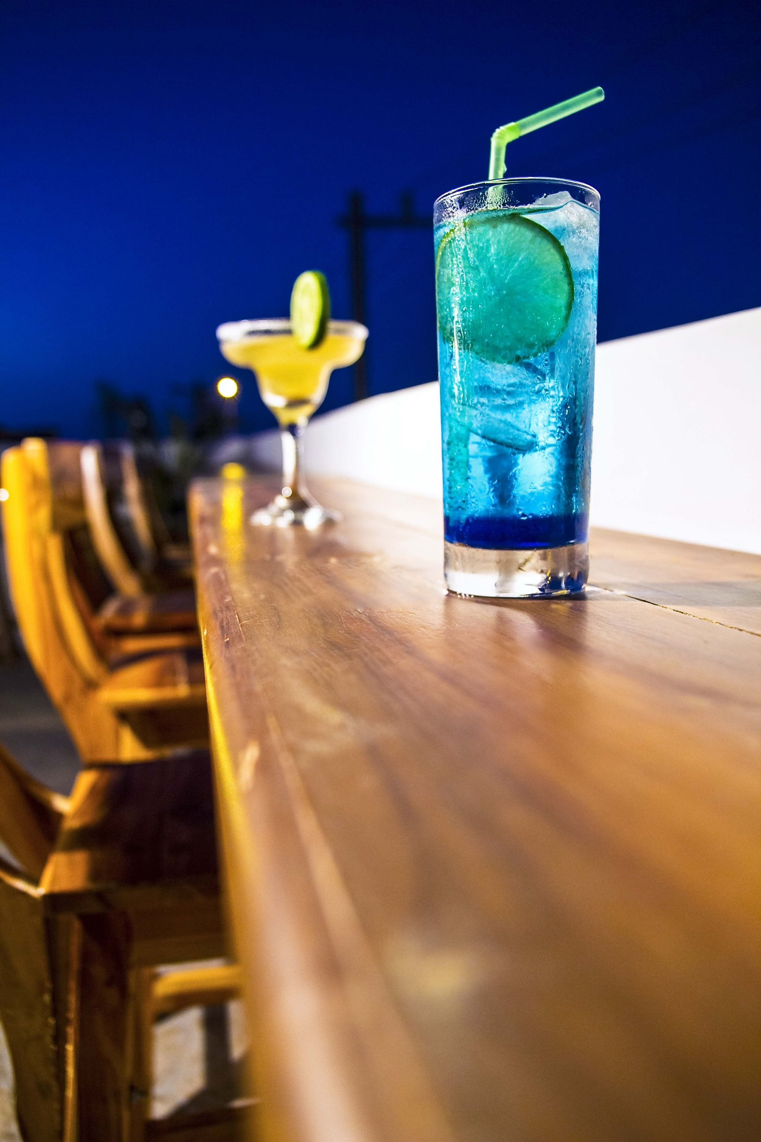 Un bar con un vaso alto de bebida azul, rodaja de lima y pajita, y un vaso más pequeño con rodaja de limón.