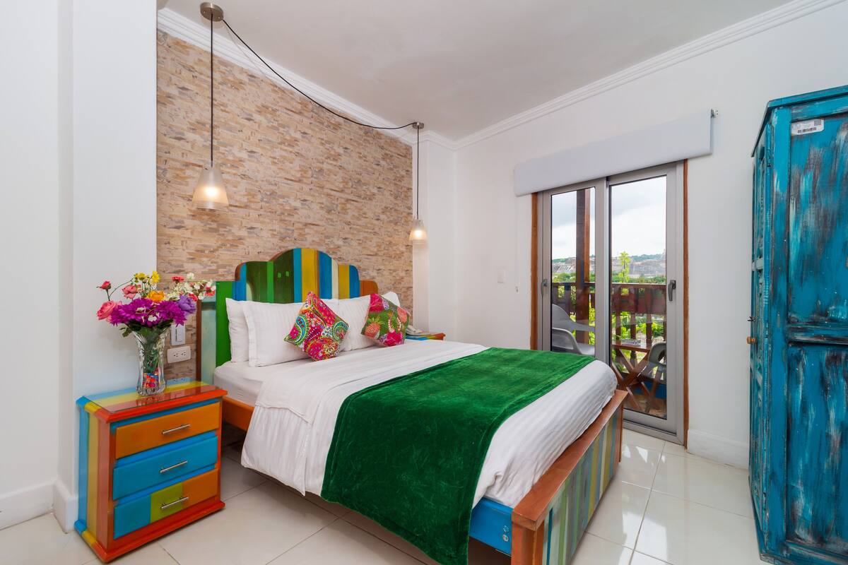 A bedroom with a brick wall, a bed with colorful pillows, a green blanket, a dresser with drawers, and a view through a sliding door.