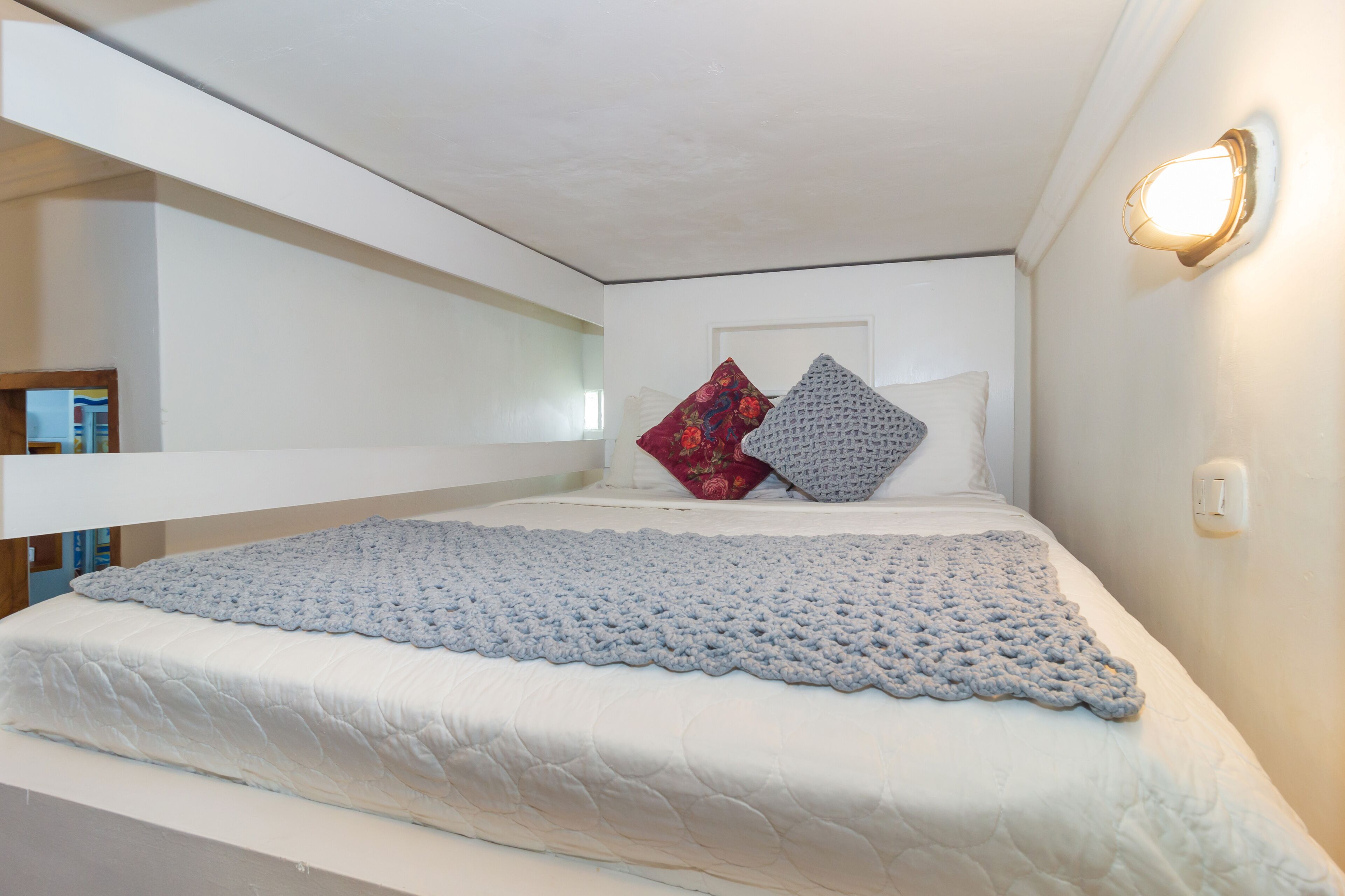 Una cama litera con una manta azul de punto, ropa de cama blanca y dos almohadas (una roja y una gris).