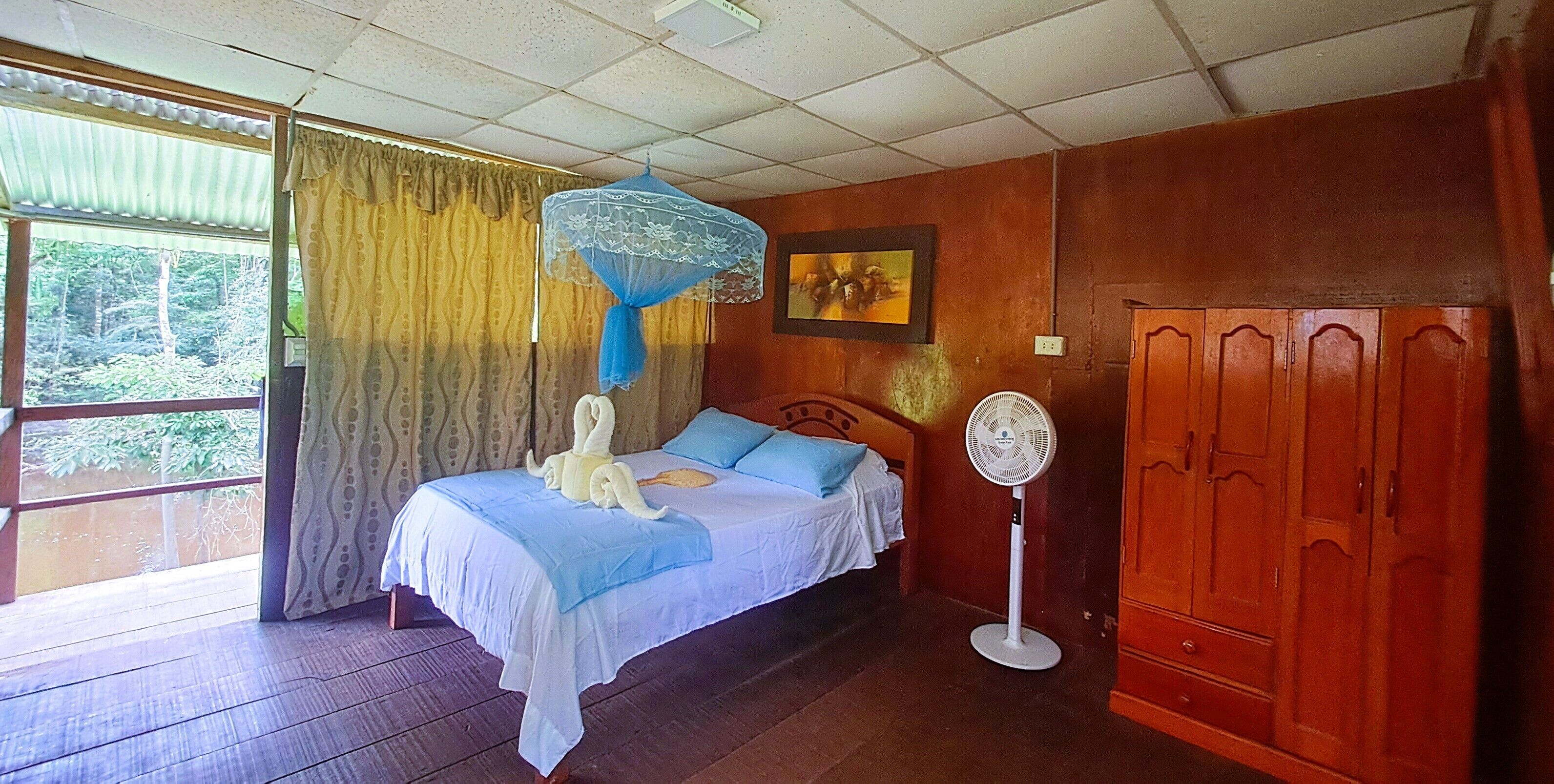 A room with a bed, a standing fan, a wardrobe, and a window with curtains.