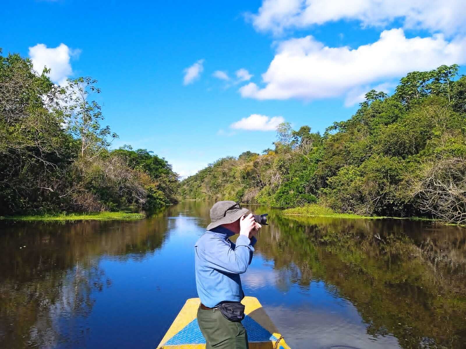 Una persona en un bote fotografiando un río tranquilo rodeado de abundante vegetación.