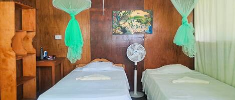 A room with two beds, a wooden shelf, a painting, and a standing fan.