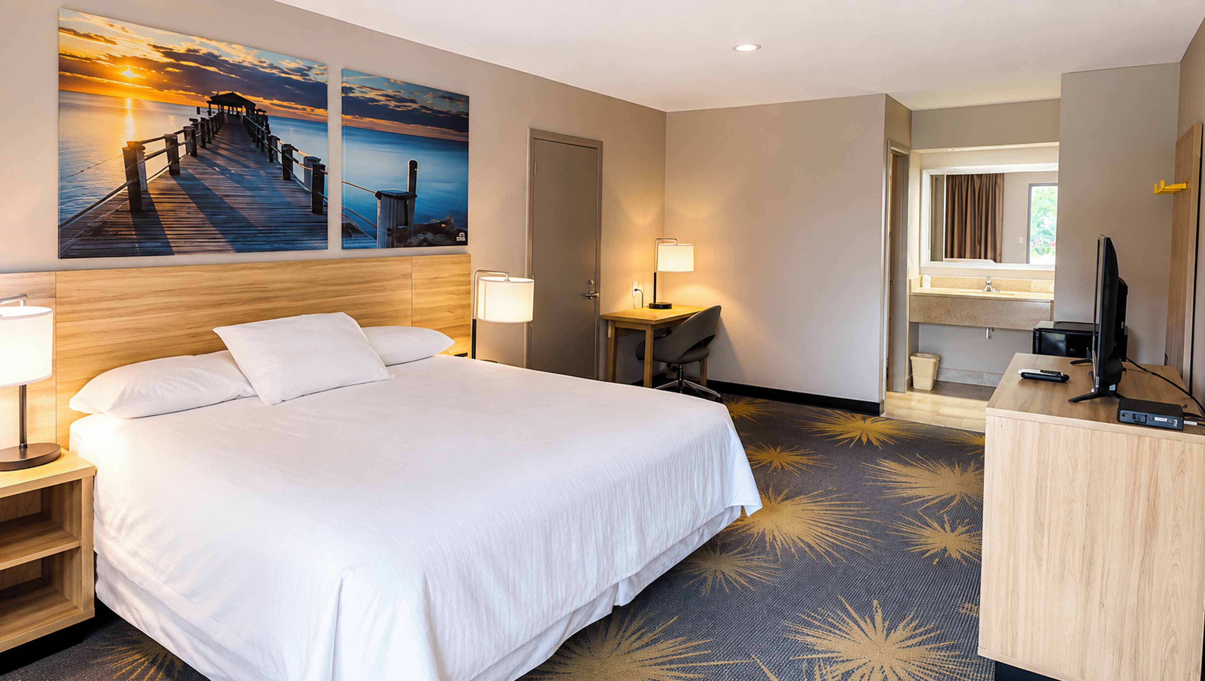 Habitación de hotel con una cama grande, un escritorio, una silla, un televisor y un baño con espejo y lavabo.