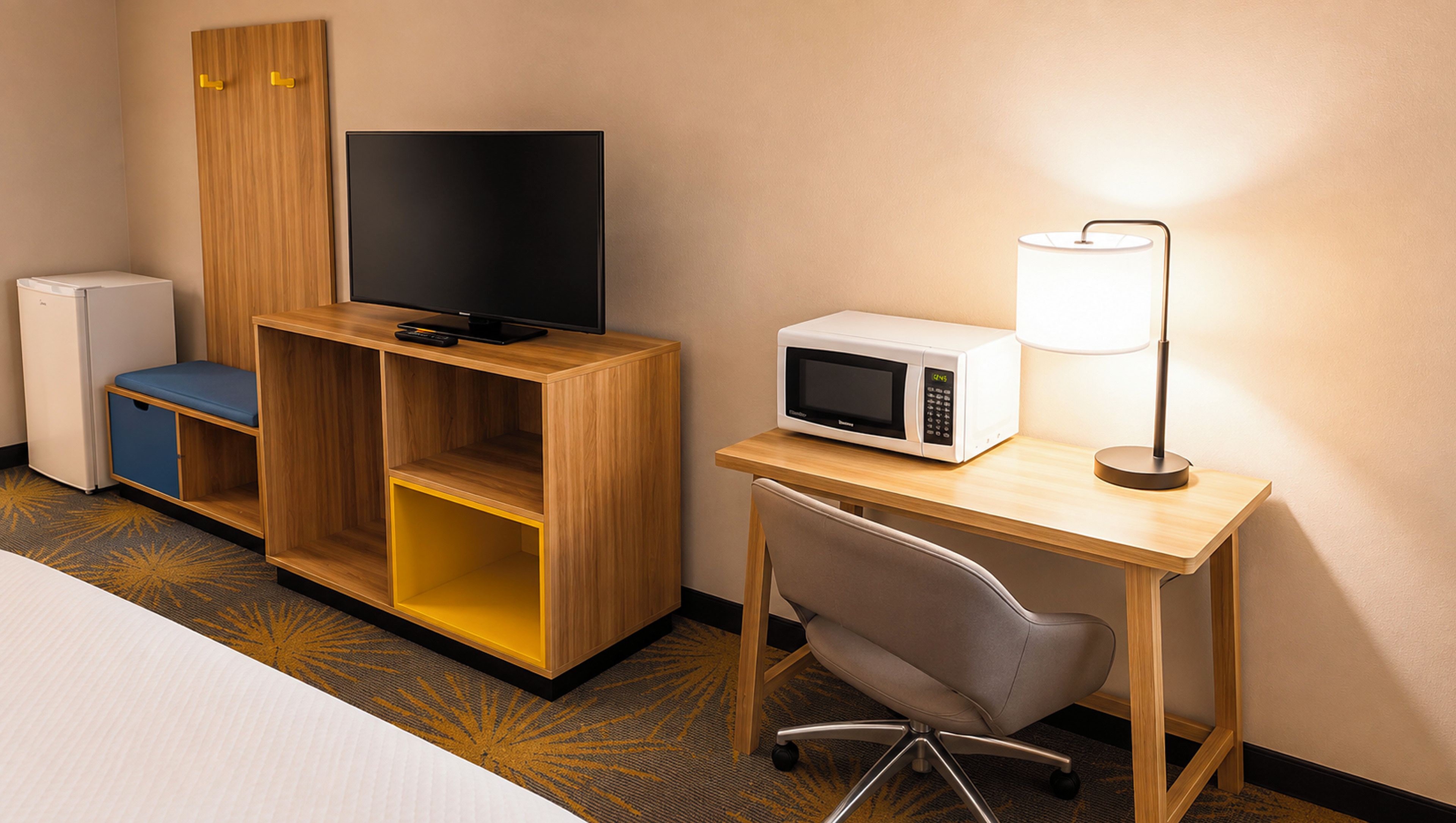 Habitación de hotel con mueble de entretenimiento de madera, televisor, microondas, escritorio con lámpara y una silla.