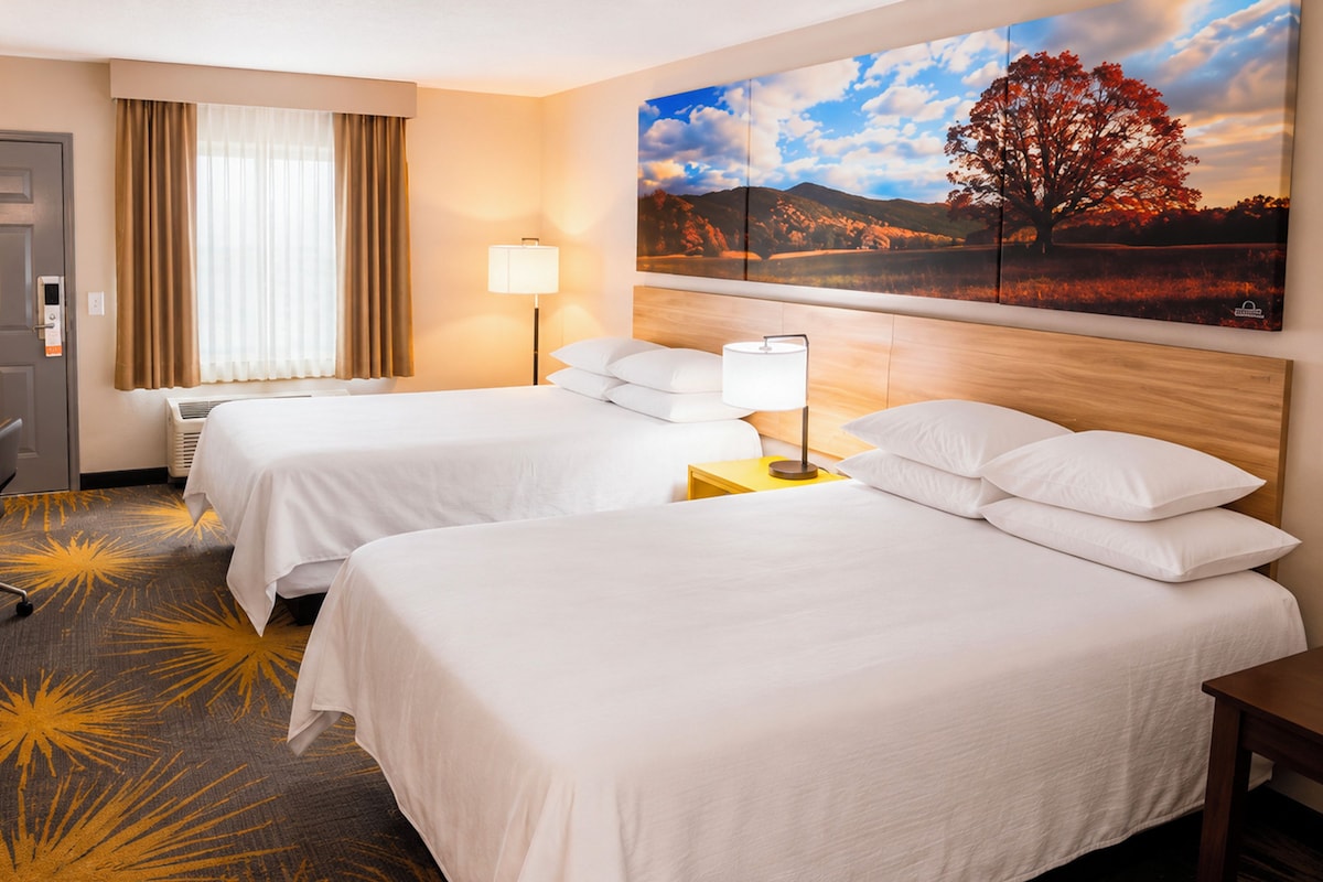 A hotel room with two beds, a desk, a TV, and a painting of a tree.