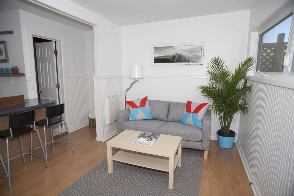 A living room with a grey sofa, a wooden coffee table, a lamp, a potted plant, and a dining area with a table and chairs.