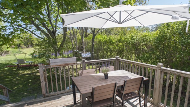 A wooden deck with a table and chairs, surrounded by a wooden railing, overlooking a grassy area with trees and a hammock.