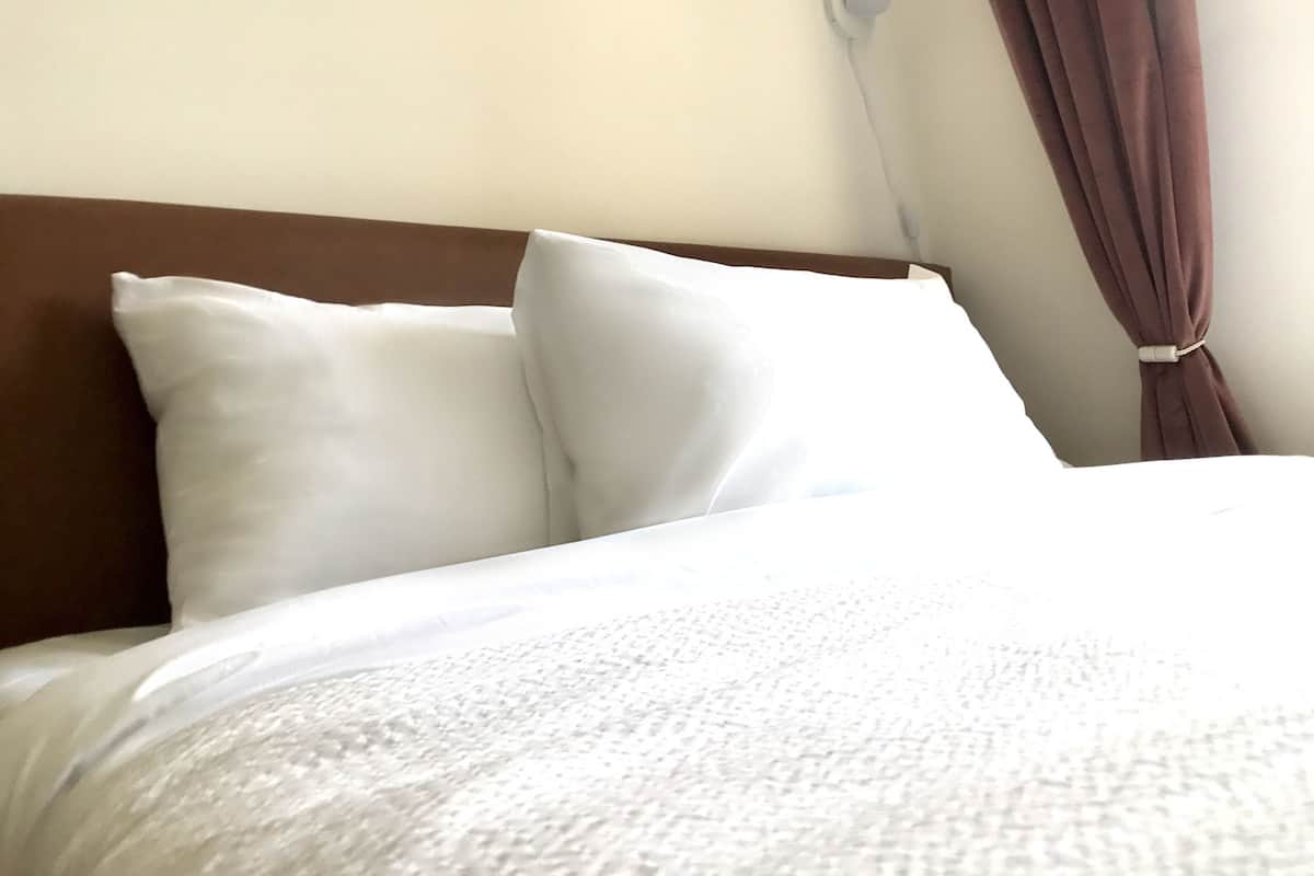 Ein ordentlich bezogenes Bett mit weißer Bettwäsche und Kissen, ein braunes Kopfteil und ein Fenster mit braunen Vorhängen.