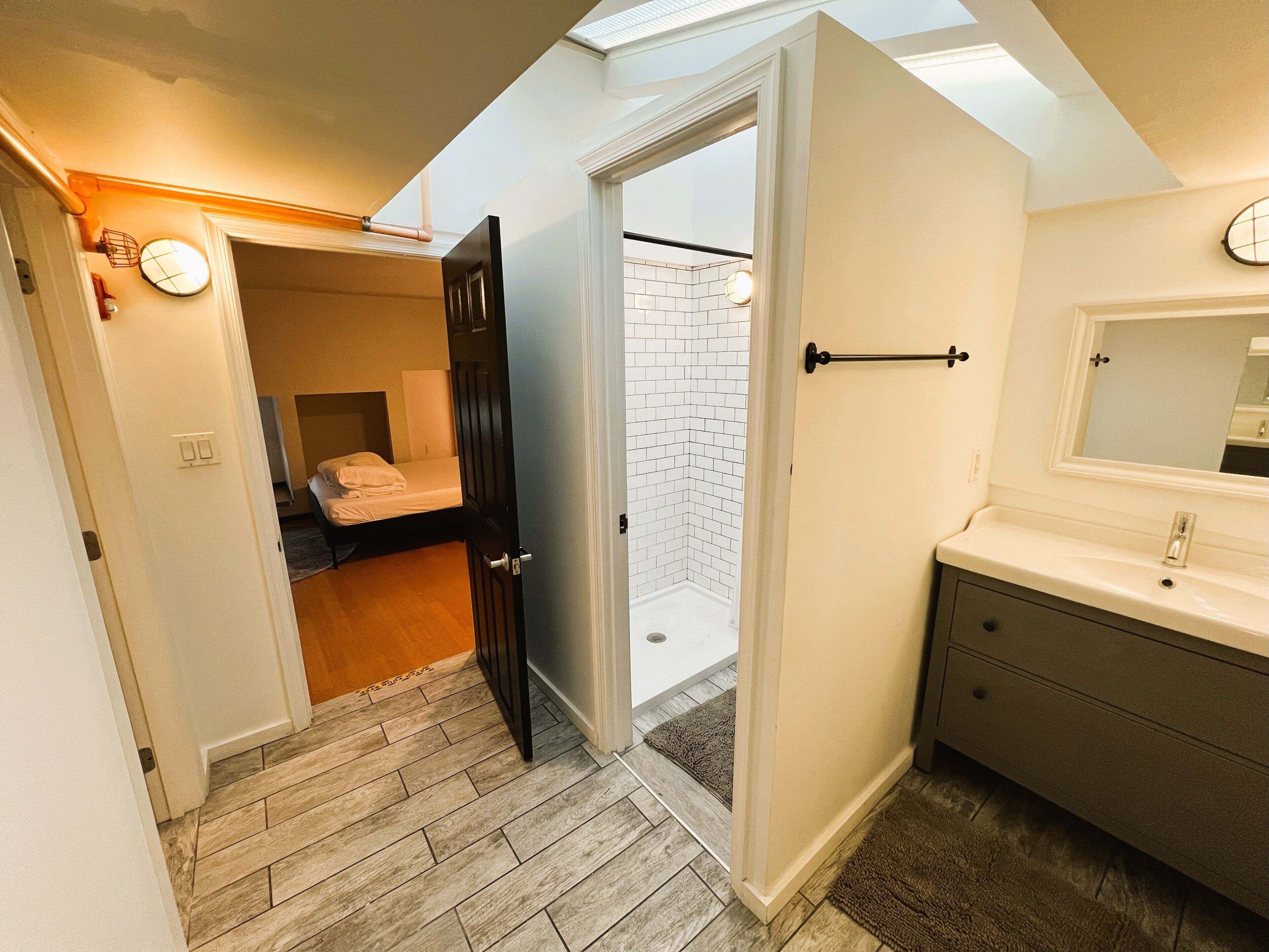 Un dormitorio compacto con cama, un baño pequeño con lavamanos y espejo, y vista al exterior por una puerta.