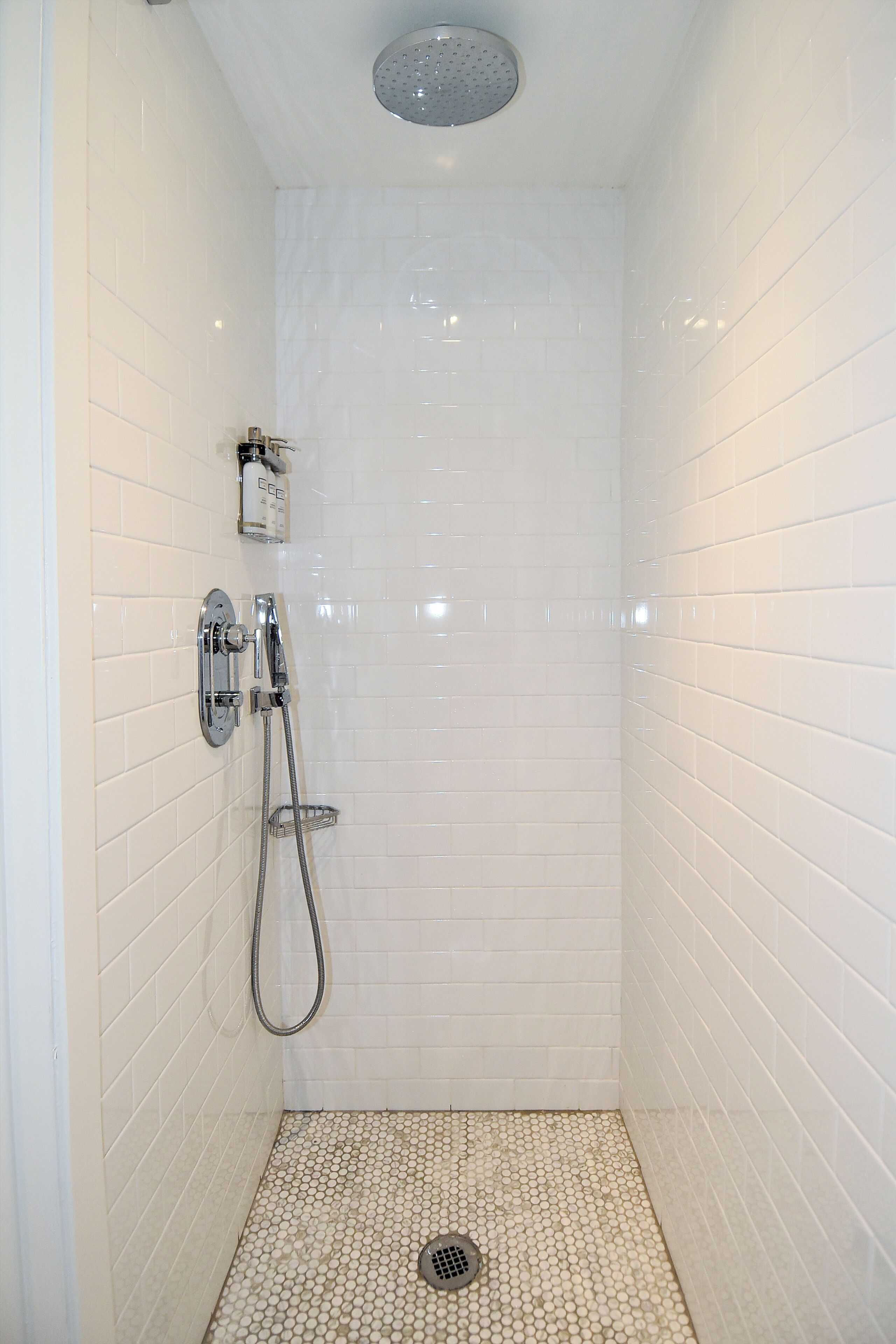 A small, white-tiled shower room with a showerhead, handheld shower, and a wall-mounted faucet.