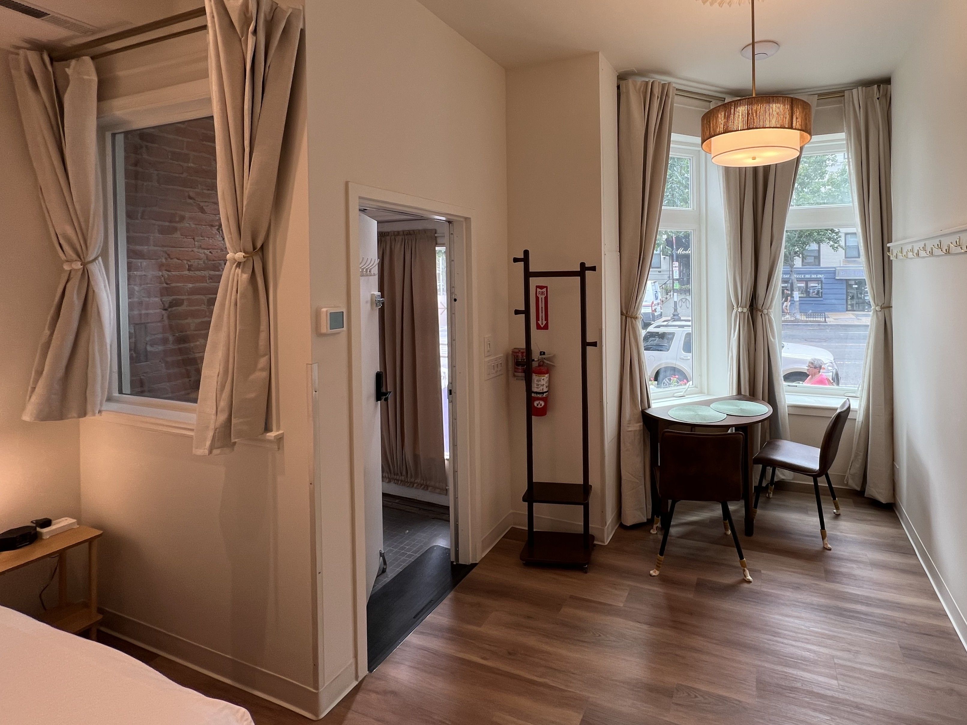Een hotelkamer met een houten vloer, een bed, een bureau met stoel, een raam met gordijnen en een hanglamp.
