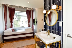 A bedroom with a bed, a window with curtains, a bedside table, and a bathroom with a sink, mirror, and tiled walls.