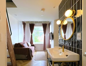 A bedroom with a bed, a window with curtains, a bathroom with a sink and mirror, and a view of greenery outside.