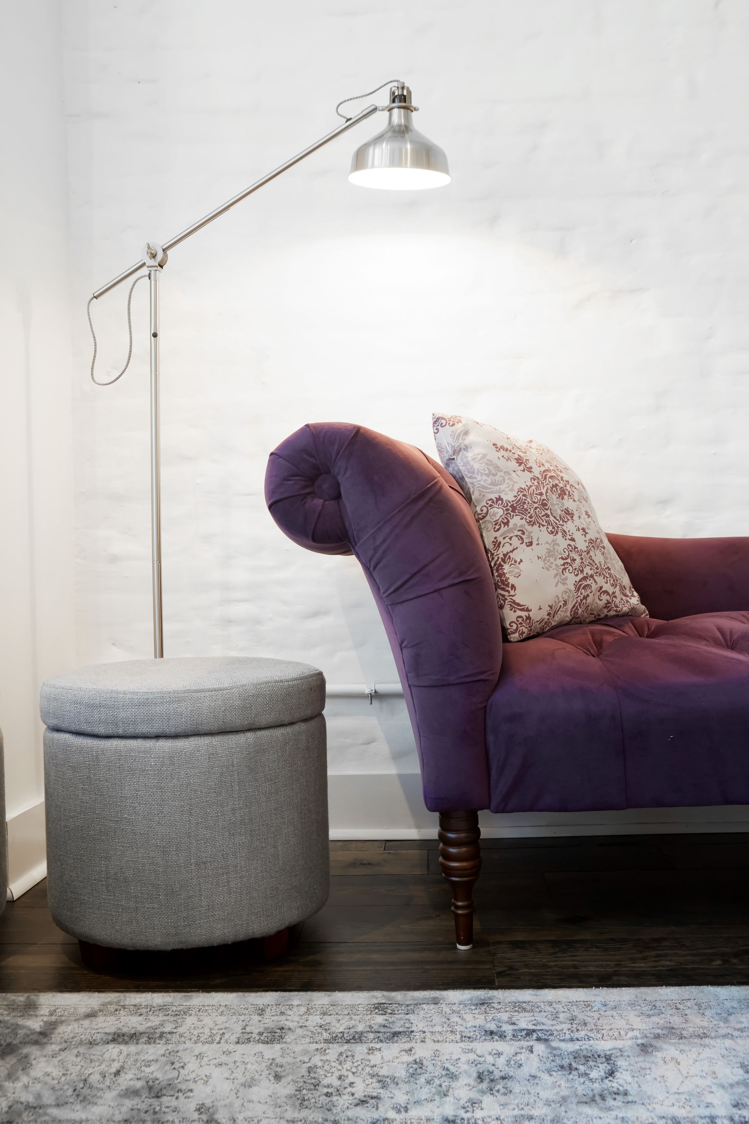 Ein violettes Sofa mit einem gemusterten Kissen, ein grauer Hocker und eine Stehlampe an einer weißen Wand.