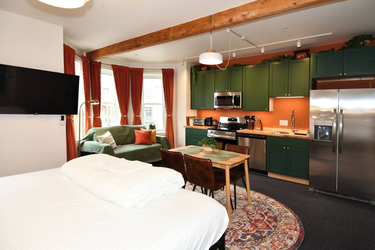 Ein modernes Hotelzimmer mit einem großen Bett, einem Esstisch, einer Küchenzeile mit grünen Schränken, einem Flachbildfernseher und einem Fenster mit roten Vorhängen.