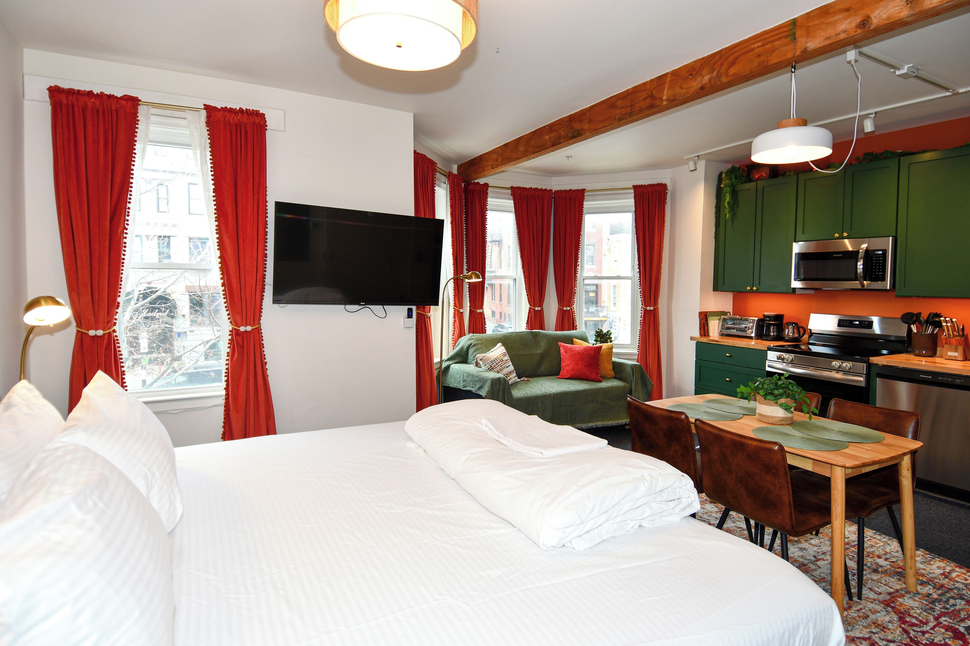 Une chambre d’hôtel comprenant un lit, une télévision, une table à manger avec des chaises, un coin cuisine avec un four à micro-ondes et une cuisinière, et des rideaux rouges.