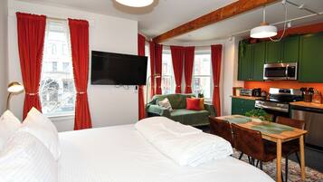 Une chambre d’hôtel comprenant un lit, une télévision, une table à manger avec des chaises, un coin cuisine avec un four à micro-ondes et une cuisinière, et des rideaux rouges.