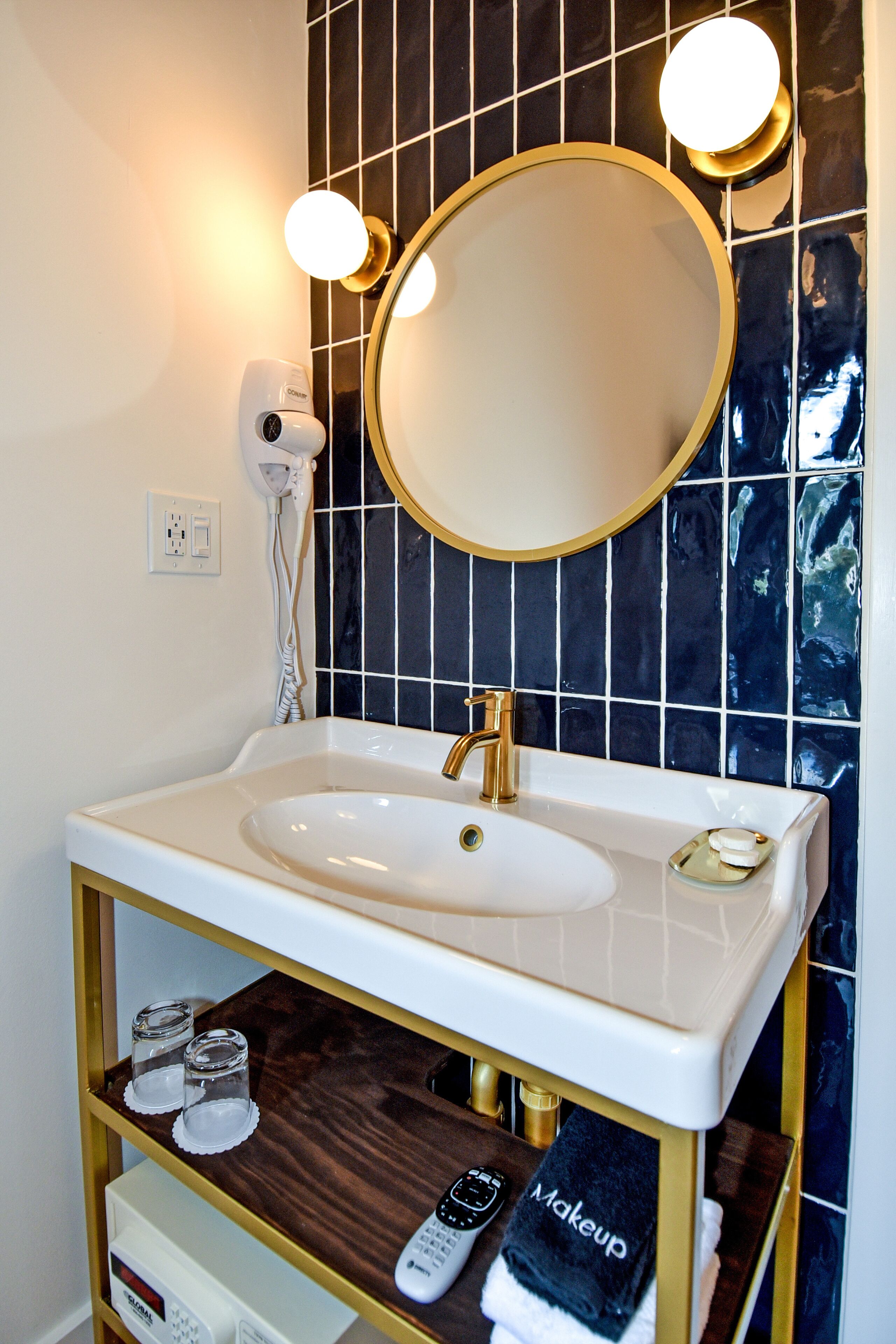 A bathroom with a white sink, gold fixtures, a round mirror, and a towel rack with a 'Makeup' towel.