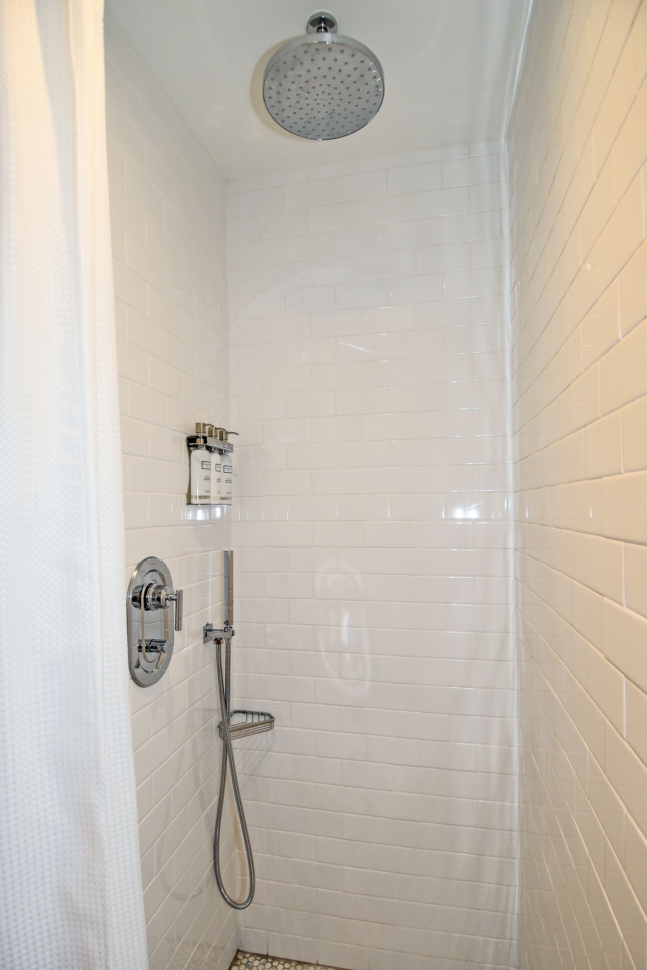 Un vestuario de ducha pequeño, con azulejos blancos, un cabezal de ducha manual y un grifo instalado en la pared.