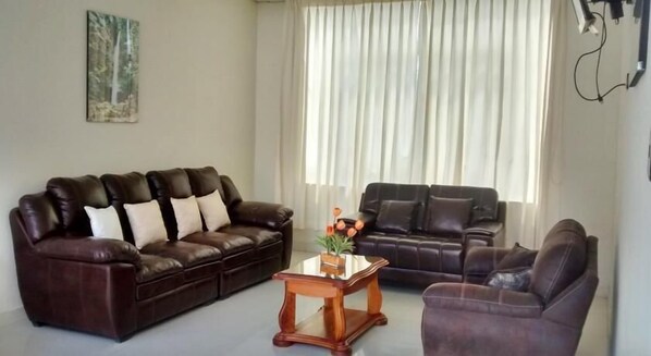 A living room with a dark brown leather sofa set, a wooden coffee table with a vase of flowers, and a large window with sheer curtains. - Royal Kerkus Hotel (Tarapoto)