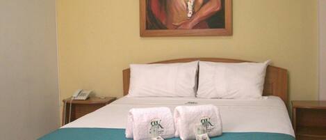 A hotel room with a wooden bed, a painting on the wall, and an air conditioner above the bed.