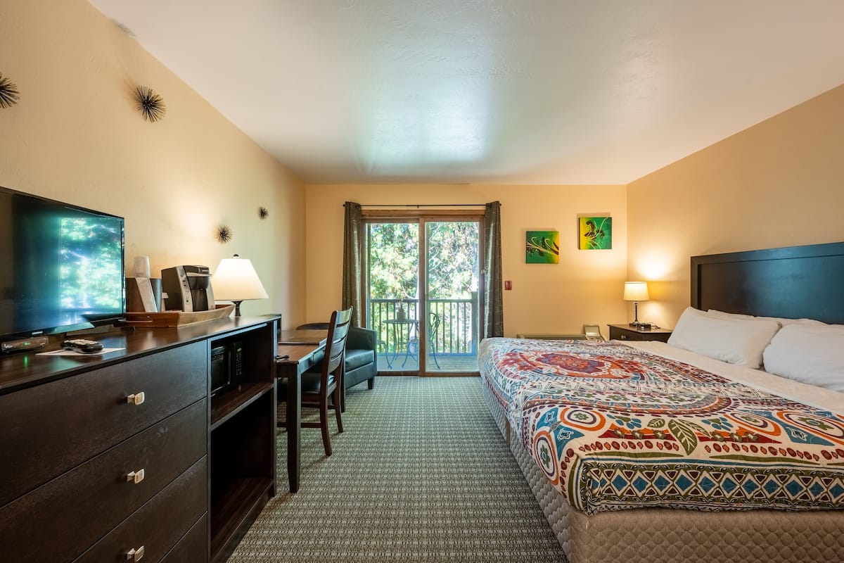 A hotel room with a bed, a desk, a television, and a balcony with a view of trees.