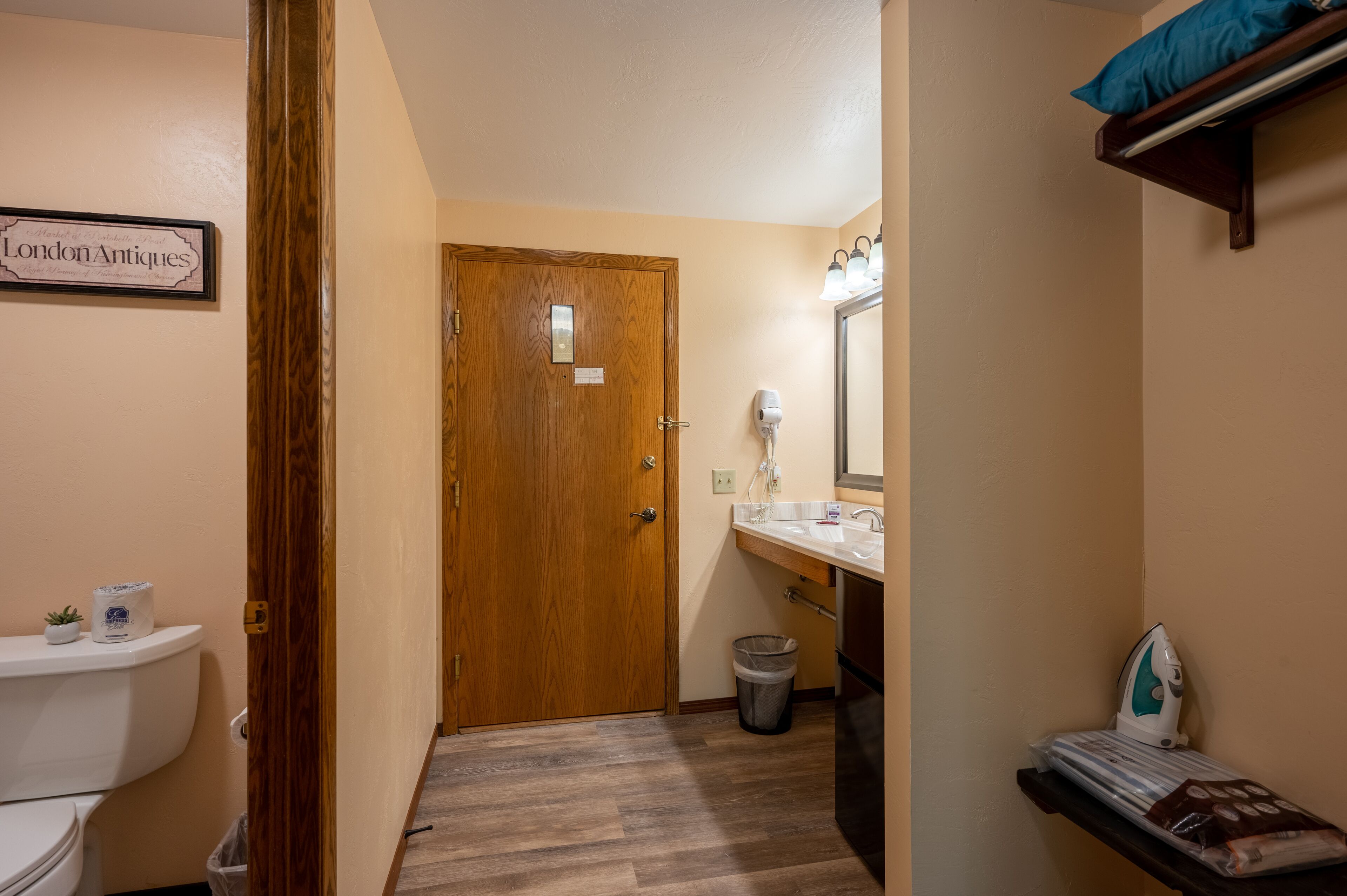 A narrow hallway with a wooden door, a white toilet, a trash can, and a bathroom with a sink and mirror.