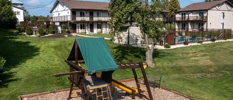 A playground with a slide and climbing structure, surrounded by a well-maintained lawn and a building with a balcony in the background.