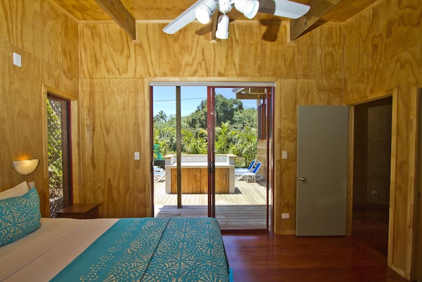 A wooden room with a bed, a ceiling fan, and a sliding glass door leading to an outdoor area with a pool and lounge chairs. - Paradise Holiday Homes Rarotonga (Rarotonga)