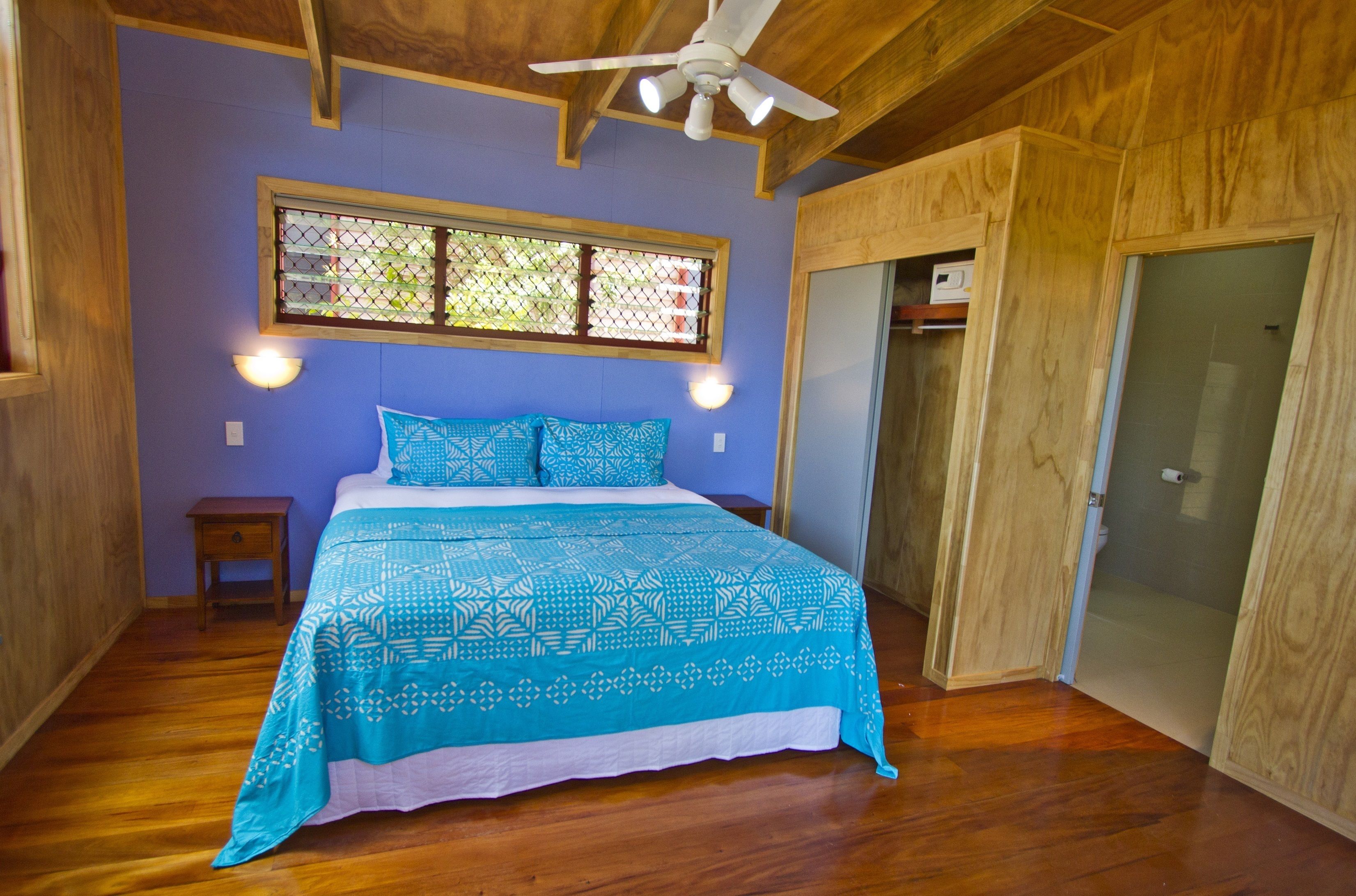 Une chambre avec un plafond en bois, un lit recouvert d’une courtepointe bleue à motifs, une fenêtre donnant sur un paysage agréable et une porte.