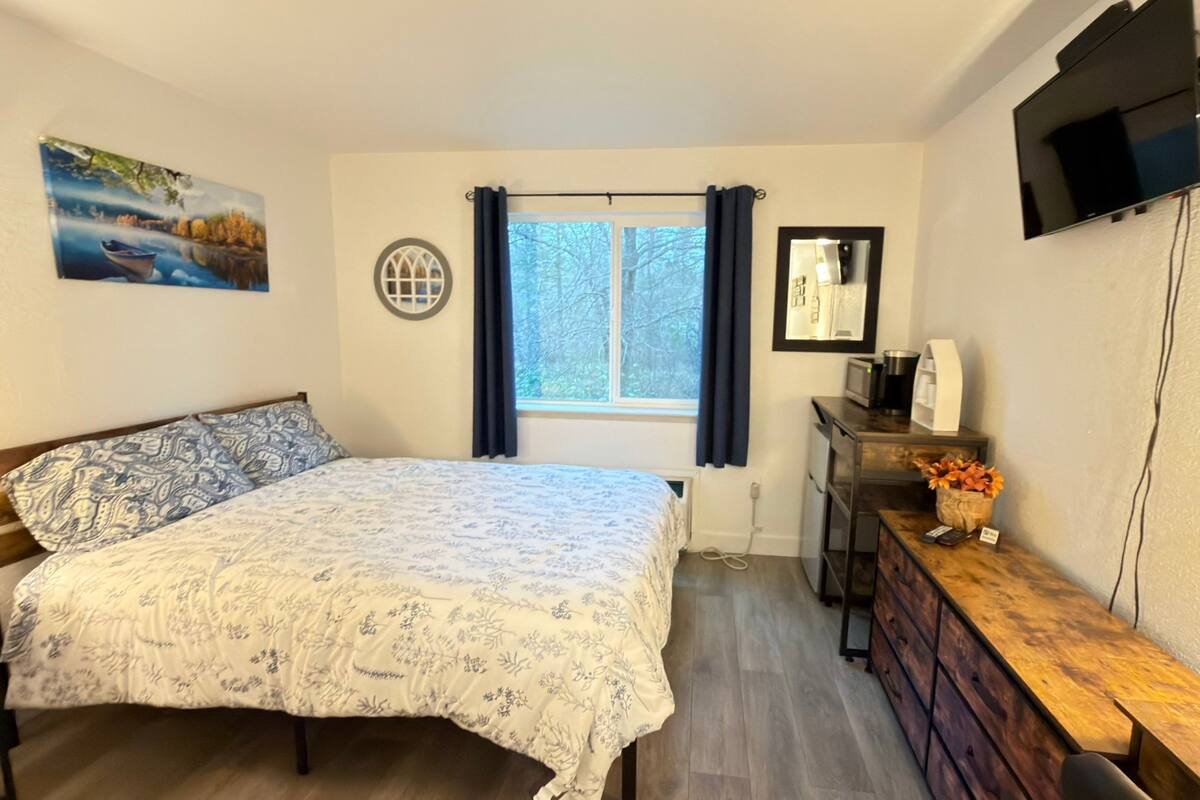 Een slaapkamer met een bed, een raam met gordijnen, een nachtkastje met een lamp en een aan de muur bevestigde televisie.