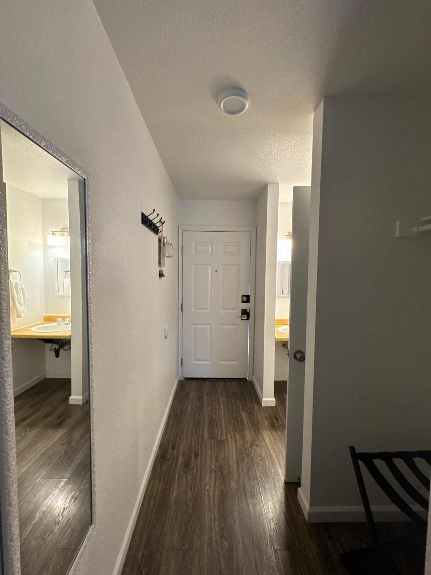 A hallway with a white door, a coat rack, and a mirror reflecting a bathroom with a sink and towel rack.