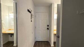 A hallway with a white door, a coat rack, and a mirror reflecting a bathroom with a sink and towel rack.